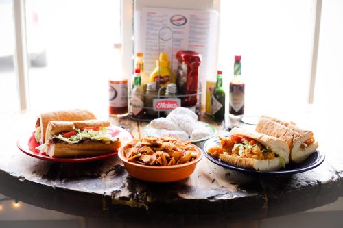 A table with two plates of sandwiches, a bowl of fries, and condiments in the background.