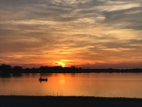 A serene sunset over a lake, with a small boat silhouetted against vibrant orange and yellow skies.