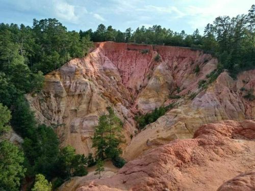 A colorful canyon with layered rock formations and green trees surrounding the area under a blue sky.
