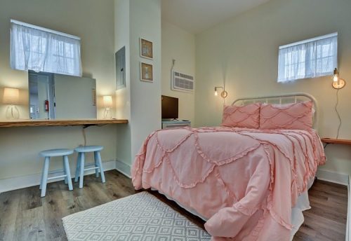 Cozy bedroom with a pink bedspread, small desk, two stools, and soft lighting from wall sconces and lamps.