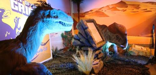 Two dinosaur models in a desert-themed exhibit, with informational signs and desert plants in the background.
