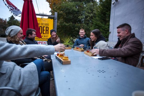A group of friends enjoying drinks at an outdoor table near a cider house, surrounded by trees and autumn colors.