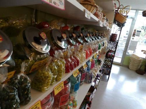 A colorful display of various candies in jars and bags on shelves in a bright store. Baskets are visible in the background.