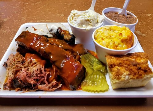 A plate of barbecue featuring ribs, pulled pork, pickles, macaroni and cheese, coleslaw, and cornbread.