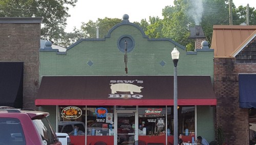 A BBQ restaurant with a green exterior and a brown awning featuring a pig logo, surrounded by trees and buildings.