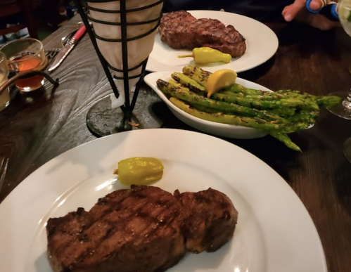 Two plates of grilled steaks with lemon and peppers, accompanied by a side of grilled asparagus on a wooden table.