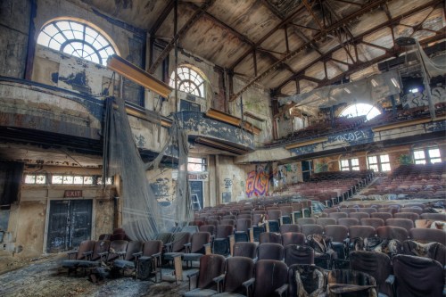 Abandoned theater interior with worn seats, peeling paint, and cobwebs, illuminated by large, dusty windows.