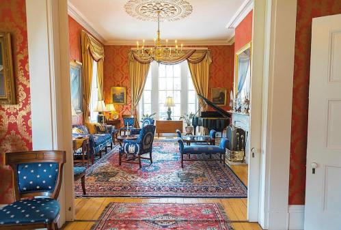 Elegant room with red wallpaper, ornate chandelier, piano, and vintage furniture, featuring a large window and decorative rugs.