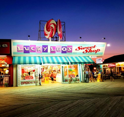 Brightly lit storefront of Lucky Leo's Sweet Shop at dusk, featuring colorful candy displays and a striped awning.