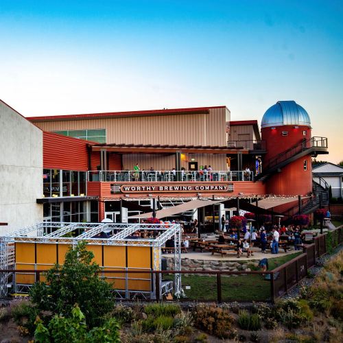 Exterior view of Worthy Brewing Company with a red observatory, outdoor seating, and people enjoying the space.