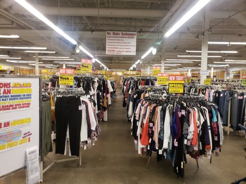 A spacious thrift store aisle filled with racks of clothing and promotional signs hanging from the ceiling.