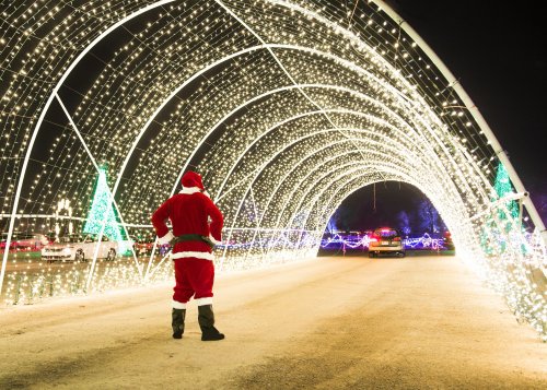A person in a Santa costume stands in front of a lit tunnel of Christmas lights, with cars driving through.