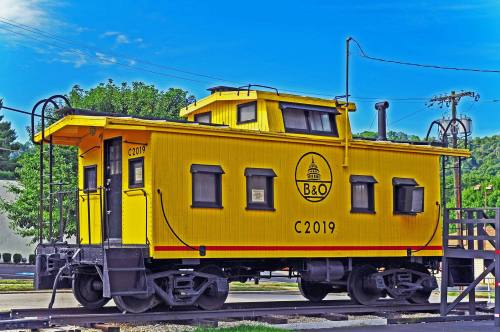 A bright yellow caboose labeled "B&O C2019" parked on tracks, surrounded by greenery and a clear blue sky.