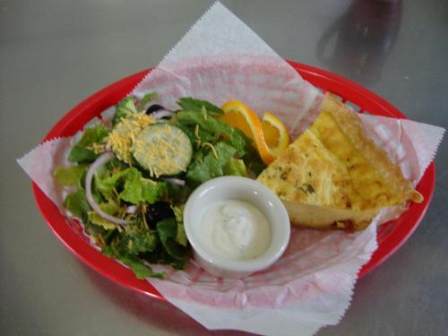 A red basket containing a fresh salad with cucumbers, broccoli, and cheese, alongside a slice of quiche and a citrus wedge.