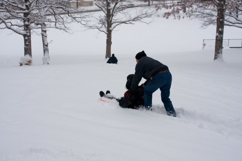 A person pulls a child on a sled through deep snow, with trees and another person in the background.
