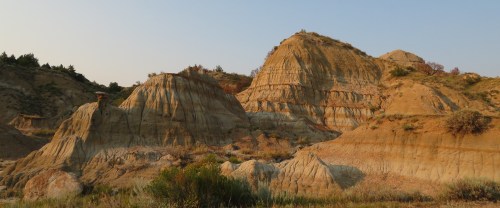 A rugged landscape featuring layered rock formations and hills under a clear sky, with sparse vegetation in the foreground.