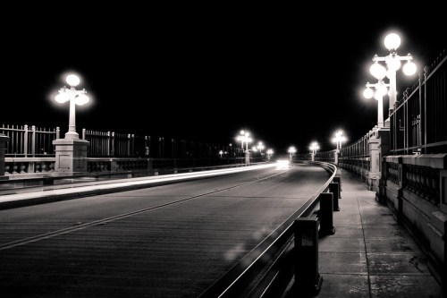 A dimly lit bridge at night, featuring ornate street lamps and light trails from passing vehicles.
