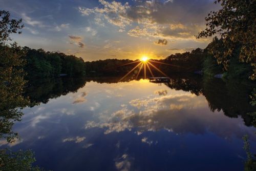 A serene lake at sunset, reflecting colorful clouds and sunlight, surrounded by lush trees.