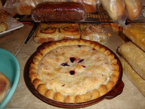 A freshly baked pie with a golden crust and berry filling, surrounded by various baked goods on a countertop.