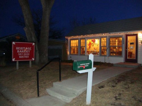 A cozy bakery with warm lights, a sign reading "Heritage Bakery," and a mailbox in front, set in the evening.