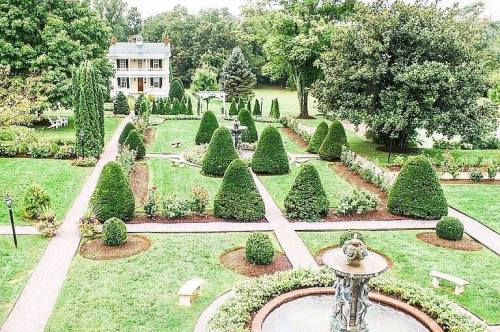 A landscaped garden featuring neatly trimmed hedges, a fountain, and a house in the background surrounded by greenery.