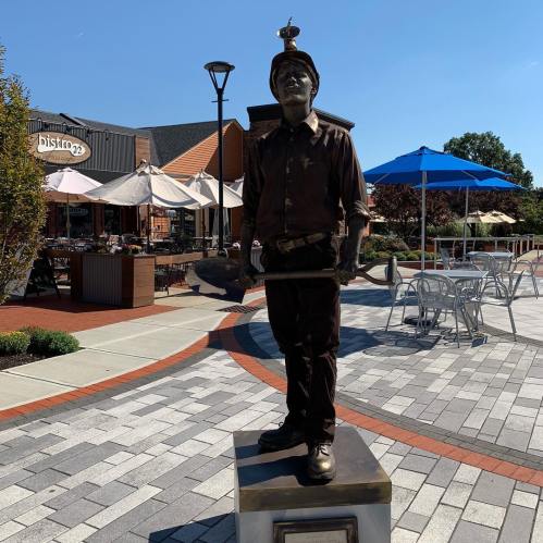 A bronze statue of a miner holding a pickaxe, standing on a pedestal in a plaza with outdoor dining areas.