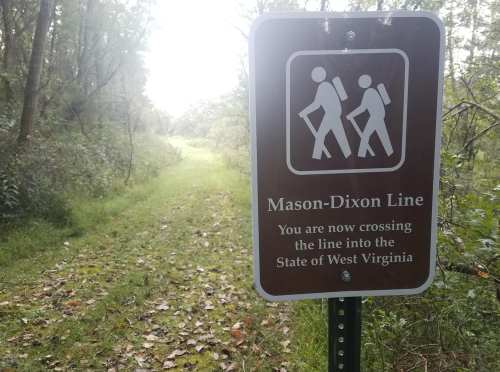 Sign marking the Mason-Dixon Line, indicating entry into West Virginia, with a wooded trail in the background.