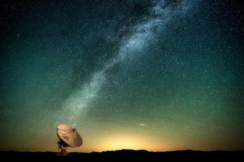 A satellite dish under a starry sky, with the Milky Way visible and a glowing horizon in the background.