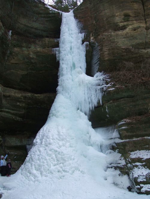 A frozen waterfall cascading down a rocky cliff, surrounded by ice and snow, with two people standing nearby.