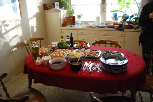 A dining table set with a red tablecloth, featuring various dishes, salads, and utensils, in a bright kitchen.