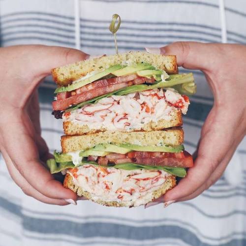 A person holds a large sandwich stacked with shrimp, lettuce, tomato, and avocado between slices of bread.