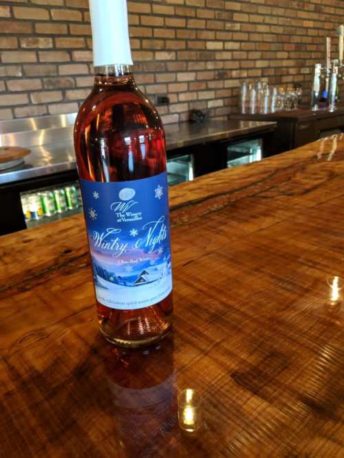 A bottle of "Wintry Nights" red wine on a wooden bar, with a brick wall and drinks in the background.