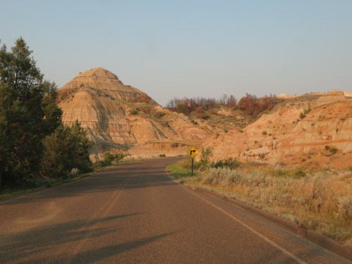 A winding road curves through colorful rock formations and hills under a clear sky. Green shrubs line the roadside.