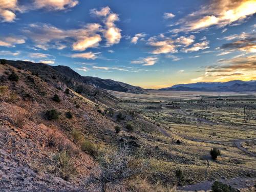 A scenic view of rolling hills and valleys under a colorful sky at sunset, with sparse vegetation and distant mountains.