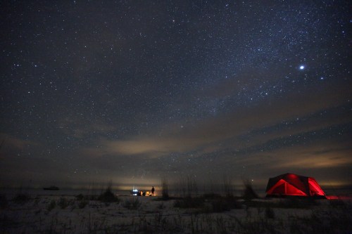 A red tent on a beach under a starry night sky, with distant lights and clouds visible on the horizon.