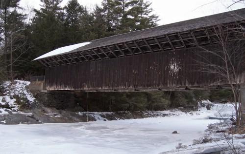 A wooden covered bridge spans a frozen river, surrounded by snow-covered trees and a winter landscape.