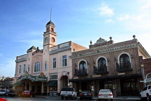 Historic theater and hotel buildings with a clock tower, set against a clear blue sky. Cars parked in front.