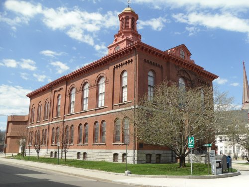 Historic red brick building with large windows and a decorative roof, surrounded by green grass and trees under a blue sky.