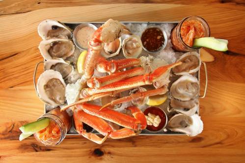 A seafood platter featuring oysters, crab legs, shrimp, sauces, and lemon, served on ice in a metal tray.