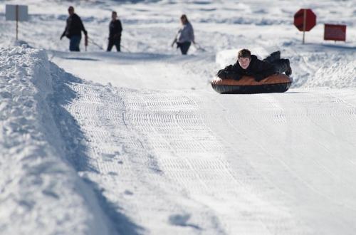 A person sleds down a snowy hill while others walk in the background, enjoying a winter day.