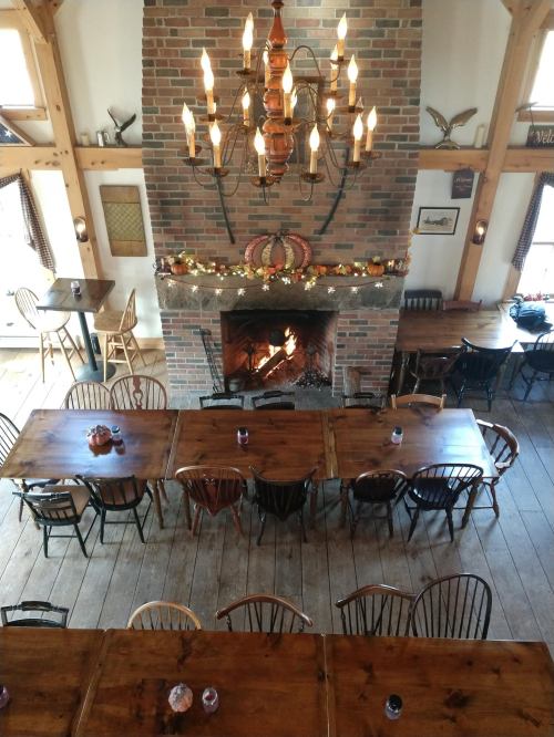 Cozy dining room with wooden tables, a fireplace, and autumn decorations, illuminated by a chandelier.