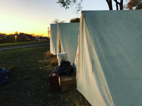 Tents set up on grass at sunset, with bags and a suitcase nearby, and a road visible in the background.