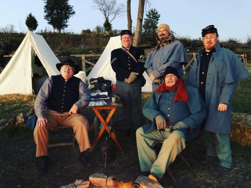 Five men in Civil War-era uniforms pose near tents and a campfire, with trees and a sunset in the background.