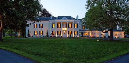A large, elegant white mansion with columns, surrounded by green grass and trees, illuminated at dusk.
