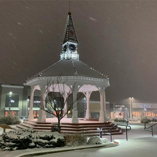 A snow-covered gazebo with a clock tower, surrounded by shops, illuminated by festive lights during a snowfall at night.