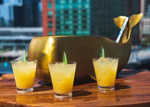 Three cocktails with yellow drinks and garnishes sit in front of a gold whale-shaped bowl on a wooden table.