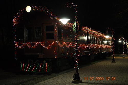 A decorated train covered in colorful lights, parked along a pathway at night, with street lamps illuminating the scene.