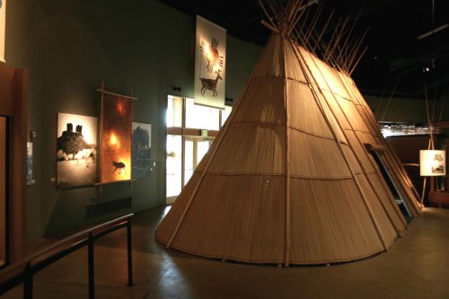 Interior view of a museum featuring a large straw teepee and art on the walls, showcasing cultural exhibits.