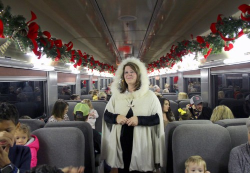 A woman in a white cape stands in a decorated train car filled with families and children during a festive event.