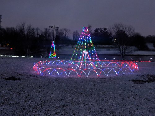 Colorful holiday lights form a large tree shape in a snowy landscape, with smaller trees in the background.